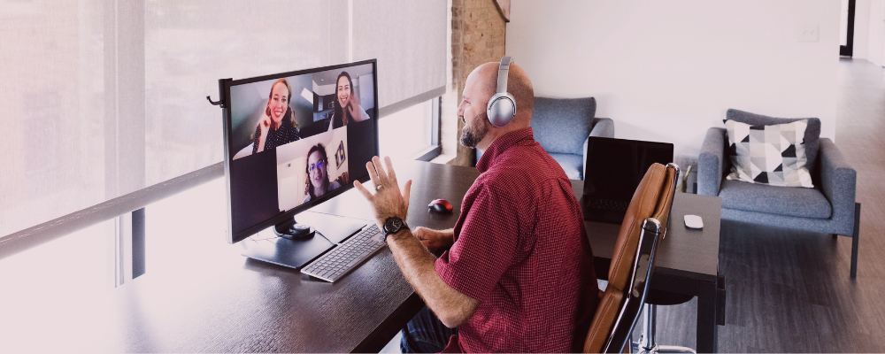 Multiple remote employees engaging in a video chat with emphasis on man seated at desk waving to the camera