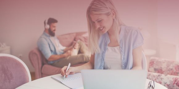 Woman smiling as she writes at a desk with man wearing headphones working behind her