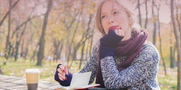 Woman wearing a scarf and drinking coffee in the park as she writes in a journal